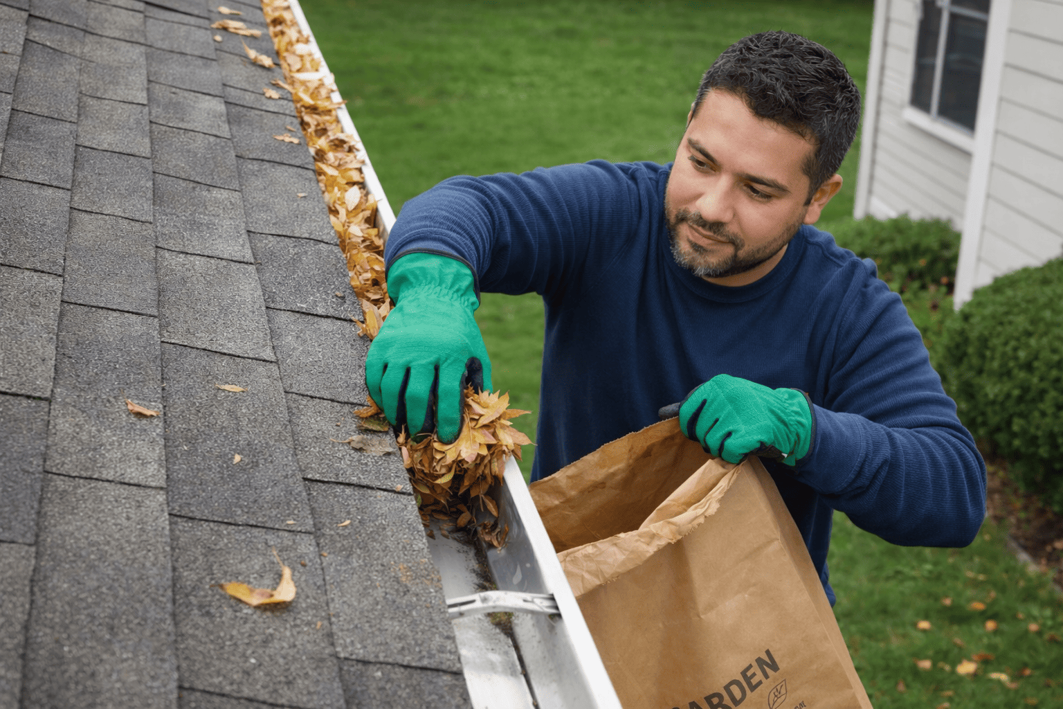 Professional gutter cleaning technician removing leaves from a residential roof gutter to prevent clogs and water damage.